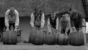Foto, zwart/wit, met ouderwets geklede mannen die grote tonnen over de grond voort rollen, op achtergrond rieten dak.