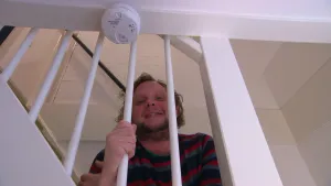 Foto van presentator die door spijlen van een trap kijkt, erboven aan balk zie je een rookmelder.