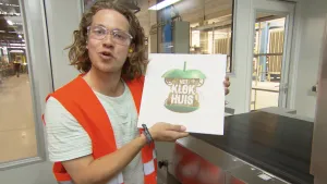 Foto van presentator ergens binnen in een (kantoor)gebouw die plaat met het logo van Klokhuis erop vasthoudt.