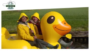 Foto van een grote nagemaakte gele badeend in het water waarin 2 personen in gele regenkleding zitten.