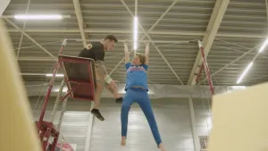 Foto van een gymnastiekzaal/sportzaal met man die op hoge bank zit aan een hoge rekstok, aan die rekstok hangt een jongetje.