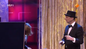 Foto van man, netjes gekleed met hoed op, op het podium, verder zie je nog deel van een vrouw met een scherm ervoor.