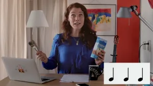 Foto van presentator aan tafel/bureau in woonkamer achter laptop, ze houdt bestek en een doosje in haar handen, rechtsonder in beeld drie muzieknoten op een rij.