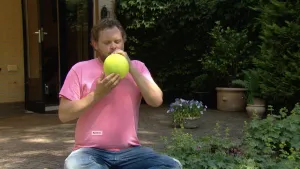 Foto van presentator die in een tuin zit met een geelgroene ballon aan zijn mond, laat 'm leeglopen in zijn mond.