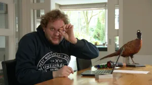 Foto van presentator die aan bureau zit met een opgezette fazant erop en een laptop, hij kijkt met één oog, één oog met zijn vingers dichtgeknepen.