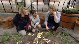 Foto van man, vrouw en meisje die geknield zitten bij aantal "vergeten" groentes die op de grond liggen in groentetuin.