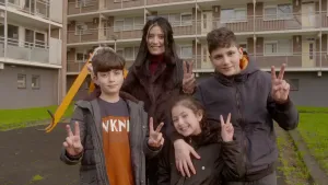 Foto van vier kinderen die buiten staan met flat op de achtergrond en laten alle vier, met hun handen, het vredesteken zien.