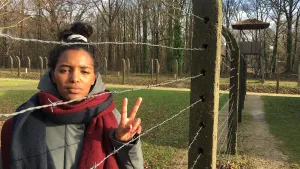 Foto van presentator achter groot prikkeldraadhek van ex-concentratiekamp die, met haar hand, het vredesteken laat zien.