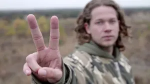 Foto van presentator buiten in de natuur die het vredesteken, met zijn hand, laat zien.