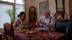 Foto van presentator die bij een oude man zit aan tafel met eten erop in huiskamer.