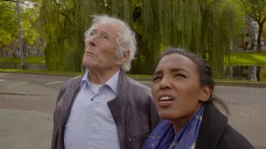 Foto van presentator die bij een oude man buiten staat op straat met op de achtergrond bos, ze kijken beiden naar iets wat niet in beeld is.