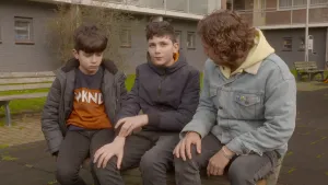 Foto met presentator die met twee jongetjes op een bank buiten zit, op de achtergrond een flatgebouw.