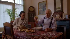 Foto van presentator en oude man die aan tafel met eten erop zitten in huiskamer.