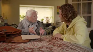 Foto van presentator die zit bij een oude vrouw aan tafel in de huiskamer.