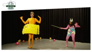 Foto van scène van toneelvoorstelling waarbij je apart geklede man en vrouw ziet, de vrouw houdt een zwaard vast, op de grond liggen balletjes.