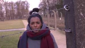 Foto van het (ex)concentratiekamp van Vught, je ziet een donkere vrouw achter een hek van prikkeldraad, op de foto verder alleen het hek en grasvelden.