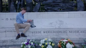 Foto van man op bank van oorlogsmonument met bloemen ervoor van de Tweede Wereldoorlog.