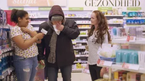 Foto van twee vrouwen in de supermarkt/drogist en een dik geklede man die zijn neus snuit in een zakdoek.