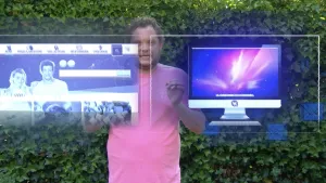 Foto van presentator voor hoge heg met in beeld ook voor hem twee grote computerschermen met van alles erop.