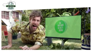 Foto van presentator die op het gras ligt in een tuin met een groot groen gekleurd computerscherm en je ziet nog deel van een groene gieter.