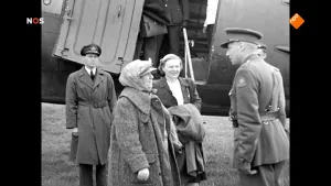 Foto van het NOS-Journaal met erop foto, zwart/wit, van Koningin Wilhelmina en aantal andere personen bij een vliegtuig, ten tijde van de Bevrijding.