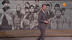 Foto van het NOS-Journaal met nieuwslezer die staat voor een heel grote zwart/wit foto met mensen erop voor een prikkeldraadhek uit de tijd van de Bevrijding.