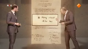 Foto van het NOS-Journaal, zwart/wit, met twee mannen die staan voor een grote afbeelding van een oude bladzijde uit 5 mei 1945.