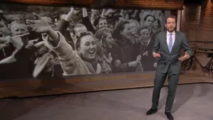 Foto van NOS-Journaal met nieuwslezer die staat voor een heel grote oude foto, zwart/wit, met veel feestvierende mensen erop tijdens de Bevrijding.