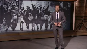 Foto van NOS-Journaal van nieuwslezer die staat voor een heel grote oude foto, zwart/wit, met feestvierende mensen op straat tijdens de Bevrijding.