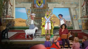 Foto van iemand die voorleest aan kinderen met op de achtergrond een decor uit een bijbelverhaal.