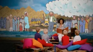 Foto van iemand die voorleest aan kinderen met op de achtergrond een decor uit een bijbelverhaal.