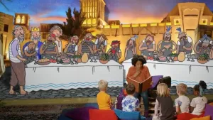 Foto van presentator die aan kinderen voorleest, zitten op het toneel, met erachter schildering van personen, profeten, die aan tafels zitten.