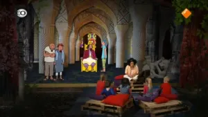 Foto van presentator die een verhaal vertelt aan kinderen die op het toneel zitten, op achtergrond decor van een kasteelzaal met personen en een koning.