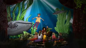 Foto van presentator die een verhaal vertelt aan kinderen die op het toneel zitten, op achtergrond decor met een walvis, een geschilderde man tussen waterplanten.