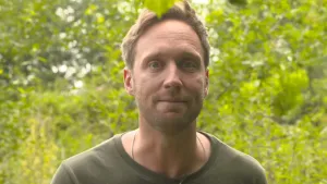 Foto van presentator, alleen zijn gezicht en schouders, met groene bomen op de achtergrond.