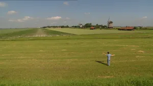 Foto van vrij hoog af van een deel van de Beemster met rechte weilanden, een persoon die er staat en twee molens in de verte.