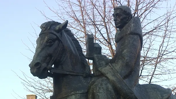 Foto van een beeld met Willibrord op een paard met boom op de achtergrond.