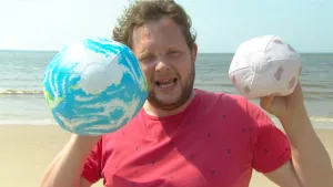 Foto van presentator op het strand bij de zee die twee ballen in zijn handen heeft, een blauwe, de aarde, een witte, de maan.