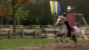 Foto van man verkleed als Middeleeuwse ridder op een rennend paard met schild en lans op renbaan.