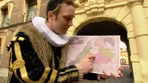 Foto van een presentator verkleed als persoon uit de zeventiende/achttiende eeuw met een kaart van Nederland van toen in zijn handen, voor een oud gebouw.