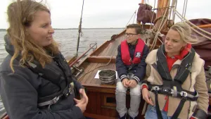 Foto van grote houten zeilboot op zee waarop aantal mensen zitten.