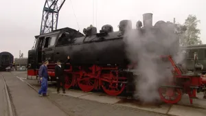Foto van een oude locomotief in werking op spoorlijn, staat nog stil, erbij staan paar personen.