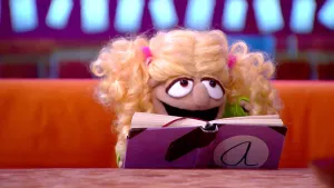 Foto van scène uit de Boterhamshow met het hoofd van Roos Maanvis (pop) die in een boek leest aan tafel.