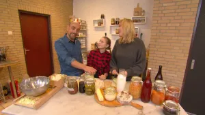Foto van een meisje, man en vrouw aan grote tafel met allerlei etenswaren erop, in weckflessen, flessen etc, in een keuken.