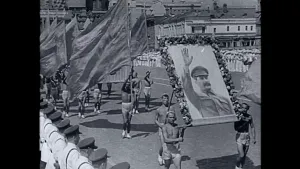 Oude foto, zwart/wit, van een militaire parade met publiek die een grote afbeelding van Jozef Stalin meedragen.
