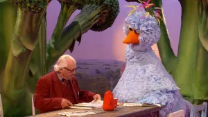 Foto van scène uit Sesamstraat waarbij Aat aan tafel zit met een boek en Pino zit erbij, op achtergrond schildering van natuurlandschap.