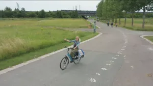 Foto van vrouw op een fiets die over een straat met grasvelden en bomen erlangs fietst.