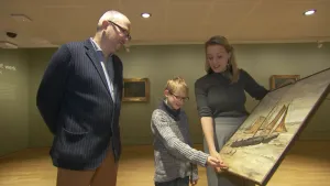 Foto van een man, kind en vrouw die een schilderij met een schip erop vasthouden/laten zien in een museum.