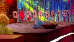 Foto van scène uit de Boterhamshow met Pi R Kwadraat aan tafel met achter hem cijfer nul tot en met negen, de één is afgedekt.