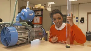 Foto van presentator in werkkamer laat een (elektro)motor zien die op tafel ligt en houdt een plastic doorzichtige buis vast.